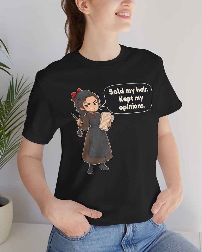 May include: A black short-sleeved t-shirt featuring a cartoon illustration of a person with a long braid, wearing a brown dress and black apron, holding a quill and papers. A speech bubble contains the text: Sold my hair. Kept my opinions.