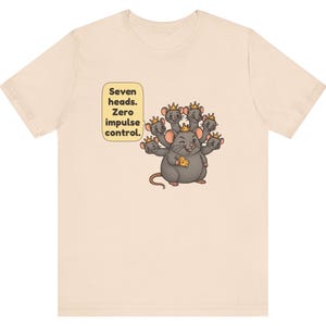 May include: A light beige t-shirt featuring a graphic of a gray seven-headed mouse wearing small gold crowns. The mouse holds a piece of yellow cheese. A speech bubble next to the mouse contains the text: Seven heads. Zero impulse control.