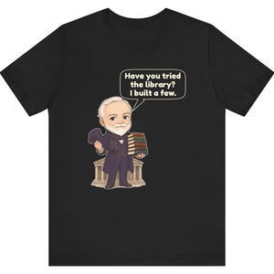 May include: A black t-shirt featuring a cartoon illustration of a bearded man in a suit holding a stack of books. A speech bubble above him contains the text, "Have you tried the library? I built a few." The man stands in front of a building.