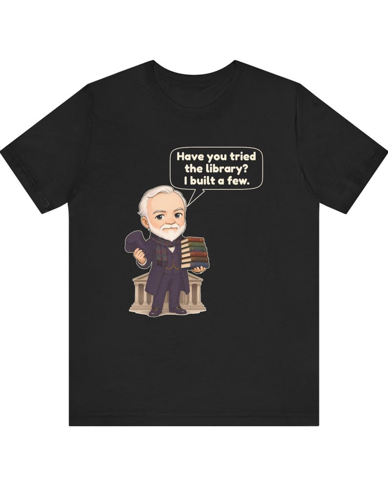May include: A black t-shirt featuring a cartoon illustration of a bearded man in a suit holding a stack of books. A speech bubble above him contains the text, "Have you tried the library? I built a few." The man stands in front of a building.