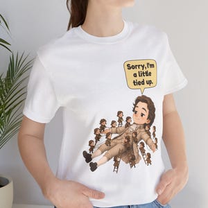 Op de afbeelding: Een wit T-shirt met korte mouwen en een afbeelding van een groot persoon in 18e-eeuwse kleding die door vele kleine figuren wordt vastgebonden. Een gele tekstballon bevat de zwarte tekst: Sorry, I'm a little tied up.