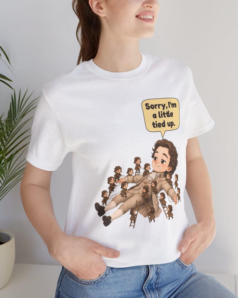 Op de afbeelding: Een wit T-shirt met korte mouwen en een afbeelding van een groot persoon in 18e-eeuwse kleding die door vele kleine figuren wordt vastgebonden. Een gele tekstballon bevat de zwarte tekst: Sorry, I'm a little tied up.