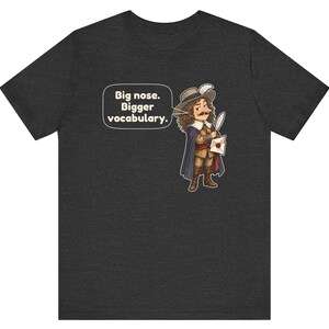 May include: A dark heather gray t-shirt featuring a cartoon illustration of a historical figure with a large nose, wearing a hat and cape, holding a quill and letter. A speech bubble contains the text: Big nose. Bigger vocabulary.