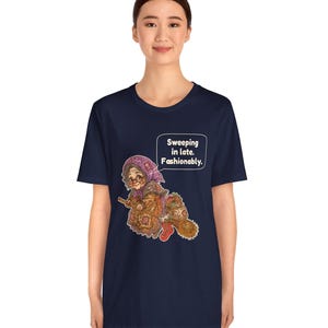 Puede incluir: Una camiseta azul marino de manga corta con un gr&aacute;fico de una persona mayor con pa&ntilde;uelo y gafas montada en una escoba. El texto en un bocadillo dice: "Sweeping in late. Fashionably.". La ilustraci&oacute;n incluye un saco y una bota roja.