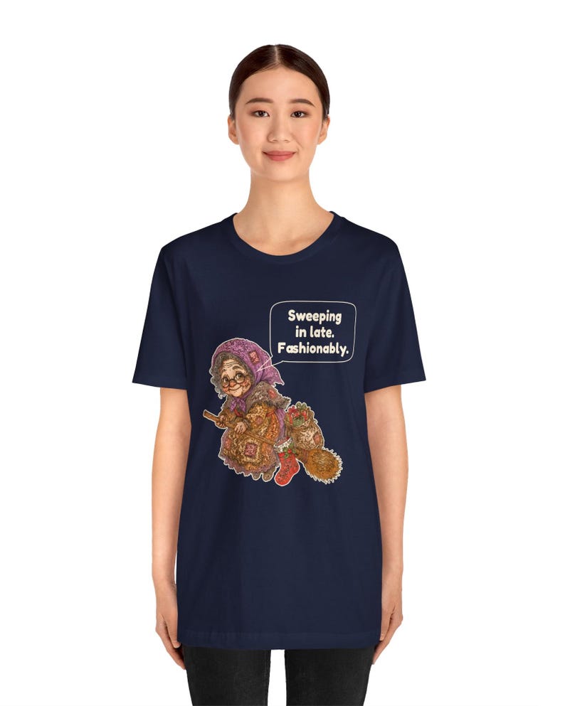 Puede incluir: Una camiseta azul marino de manga corta con un gr&aacute;fico de una persona mayor con pa&ntilde;uelo y gafas montada en una escoba. El texto en un bocadillo dice: "Sweeping in late. Fashionably.". La ilustraci&oacute;n incluye un saco y una bota roja.