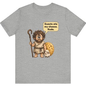 May include: A heather gray t-shirt featuring a cartoon cyclops with a brown wooden staff and a small white sheep. The cyclops stands next to a large round piece of yellow Swiss cheese. A speech bubble contains the text: Guests ate my cheese. Rude.