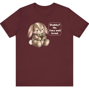 May include: A maroon short-sleeve t-shirt featuring a graphic of a brown stuffed rabbit holding holly berries. Next to the rabbit is a speech bubble with the text: Shabby? No. Very well loved. The rabbit has stitched details and a white tail.