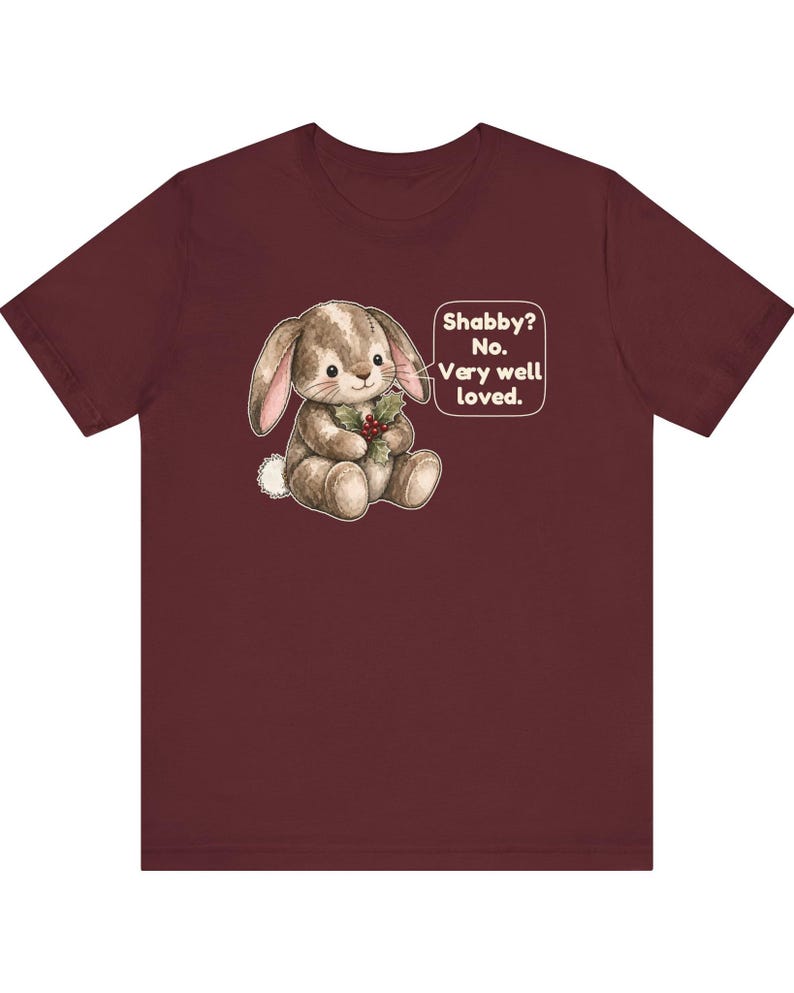 May include: A maroon short-sleeve t-shirt featuring a graphic of a brown stuffed rabbit holding holly berries. Next to the rabbit is a speech bubble with the text: Shabby? No. Very well loved. The rabbit has stitched details and a white tail.