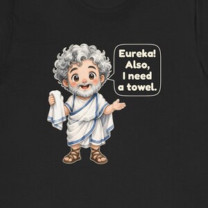 May include: A black t-shirt featuring a cartoon illustration of a bearded man with curly gray hair wearing a white toga with blue trim and brown sandals. The man holds a white towel and a speech bubble reads: Eureka! Also, I need a towel.