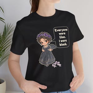 May include: A black t-shirt featuring a cartoon illustration of a person in a black dress with a purple floral crown and fan. A speech bubble contains the text: Everyone wore lilac. I wore black. A small purple ribbon is below the figure.