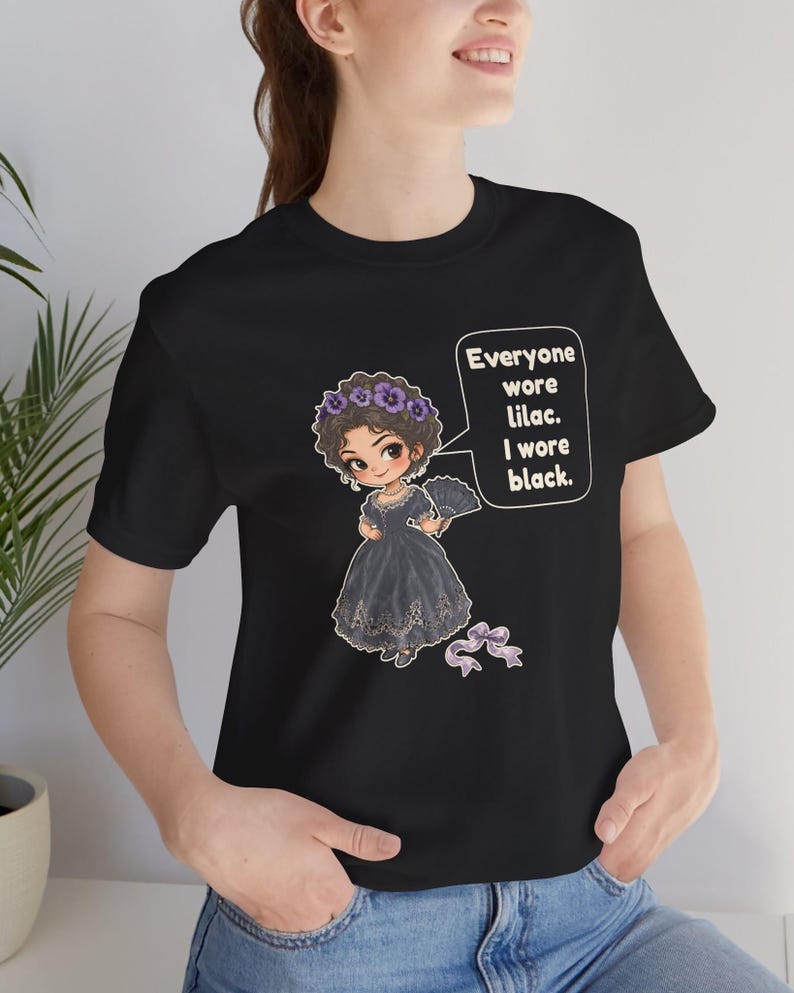May include: A black t-shirt featuring a cartoon illustration of a person in a black dress with a purple floral crown and fan. A speech bubble contains the text: Everyone wore lilac. I wore black. A small purple ribbon is below the figure.