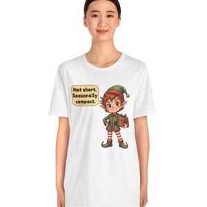 May include: A white t-shirt featuring a graphic of a cartoon elf wearing a green and red outfit with a matching hat, holding a red gift box. A yellow speech bubble next to the elf contains the black text: Not short. Seasonally compact.