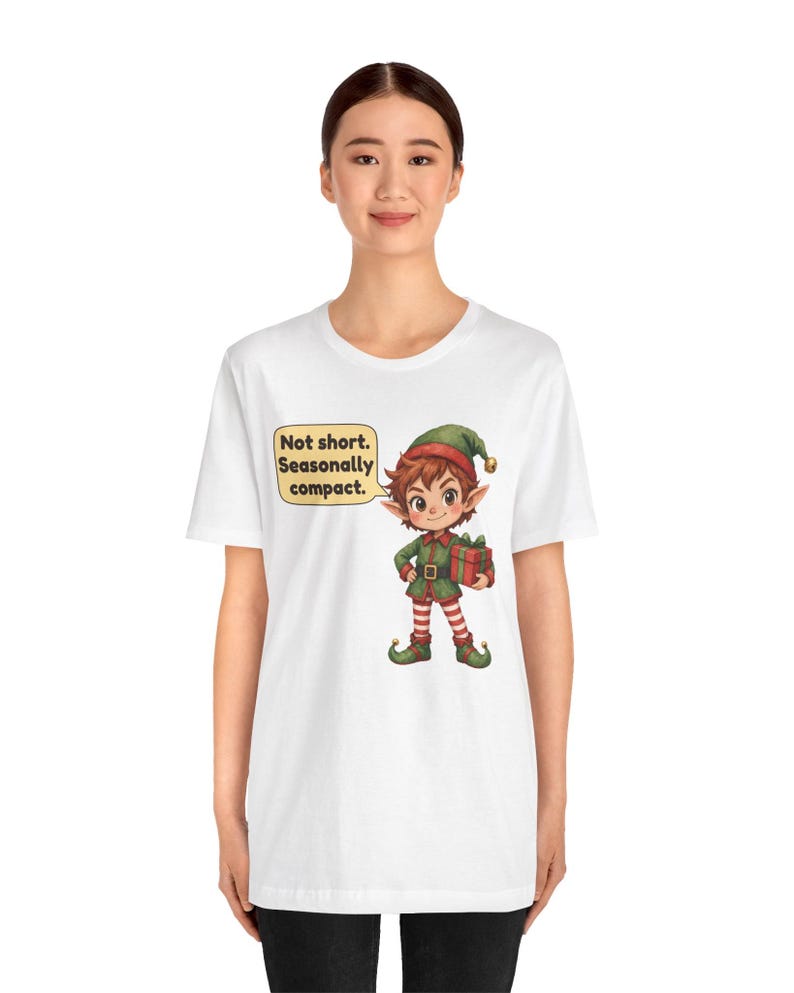 May include: A white t-shirt featuring a graphic of a cartoon elf wearing a green and red outfit with a matching hat, holding a red gift box. A yellow speech bubble next to the elf contains the black text: Not short. Seasonally compact.
