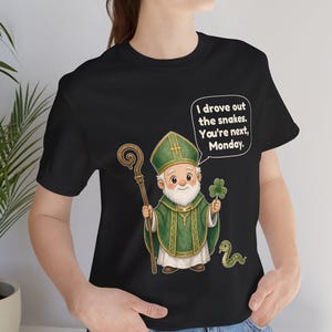 Puede incluir: Una camiseta negra con una ilustraci&oacute;n de San Patricio con vestiduras verdes y mitra, sosteniendo un bast&oacute;n de madera y un tr&eacute;bol. Una peque&ntilde;a serpiente verde est&aacute; al lado. El globo de texto dice: I drove out the snakes. You're next, Monday.