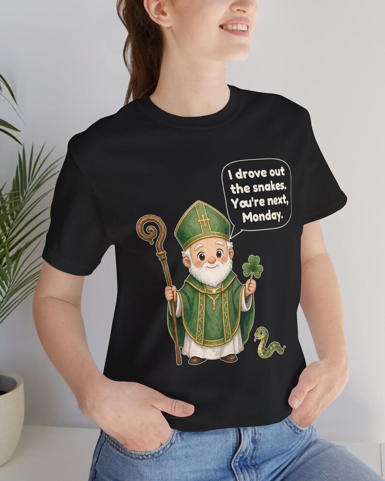 Puede incluir: Una camiseta negra con una ilustraci&oacute;n de San Patricio con vestiduras verdes y mitra, sosteniendo un bast&oacute;n de madera y un tr&eacute;bol. Una peque&ntilde;a serpiente verde est&aacute; al lado. El globo de texto dice: I drove out the snakes. You're next, Monday.