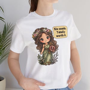 May include: A white t-shirt featuring a graphic of a cartoon character with long brown hair, a flower crown, and a green dress, holding a pomegranate. A yellow speech bubble contains the text: Six seeds. Totally worth it.