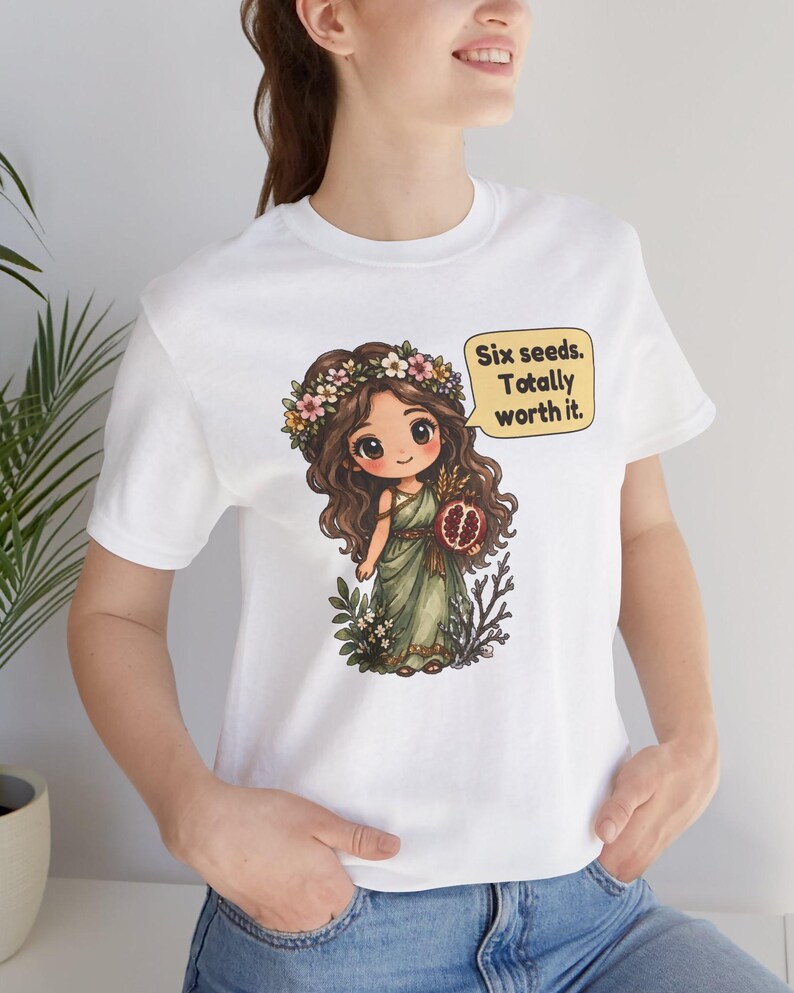 May include: A white t-shirt featuring a graphic of a cartoon character with long brown hair, a flower crown, and a green dress, holding a pomegranate. A yellow speech bubble contains the text: Six seeds. Totally worth it.