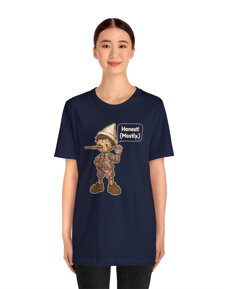 K&ouml;nnte beinhalten: Ein dunkelblaues Kurzarm-T-Shirt mit einer Grafik einer h&ouml;lzernen Marionettenfigur mit langer Nase, die einen Hut und eine kurze Hose tr&auml;gt. Eine Sprechblase neben der Figur enth&auml;lt den Text "Honest! (Mostly.)" in wei&szlig;en Buchstaben.