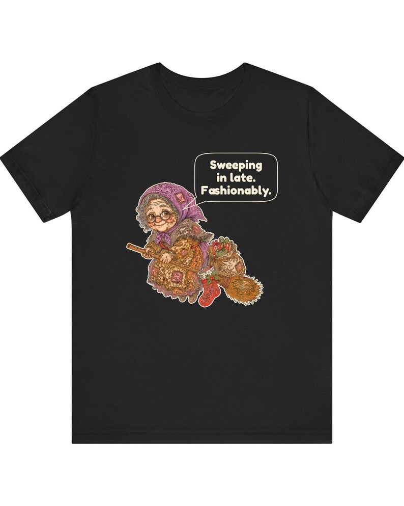 Puede incluir: Una camiseta negra con el gr&aacute;fico de una persona mayor con pa&ntilde;uelo morado y gafas montada en una escoba. La persona lleva un vestido estampado y botas rojas. Un bocadillo de texto contiene: Sweeping in late. Fashionably.