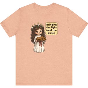 May include: A peach-colored t-shirt featuring a cartoon illustration of a figure wearing a candle crown and white robe, holding a tray of buns. A speech bubble next to the figure contains the text: Bringing the light (and the buns).