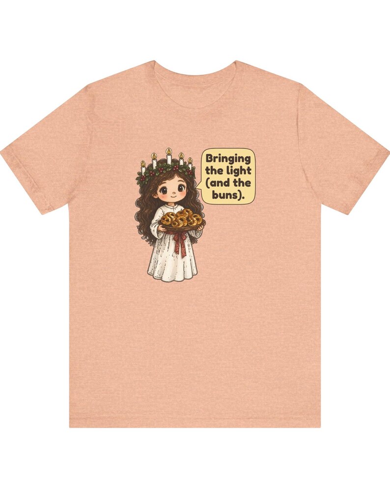 May include: A peach-colored t-shirt featuring a cartoon illustration of a figure wearing a candle crown and white robe, holding a tray of buns. A speech bubble next to the figure contains the text: Bringing the light (and the buns).