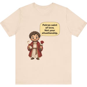 Puede incluir: Una camiseta de color beige claro con una ilustraci&oacute;n de dibujos animados de una figura barbuda con t&uacute;nica roja que sostiene un pergamino y una rosa roja. Un globo de texto contiene: Patron saint of love. Not your situationship.