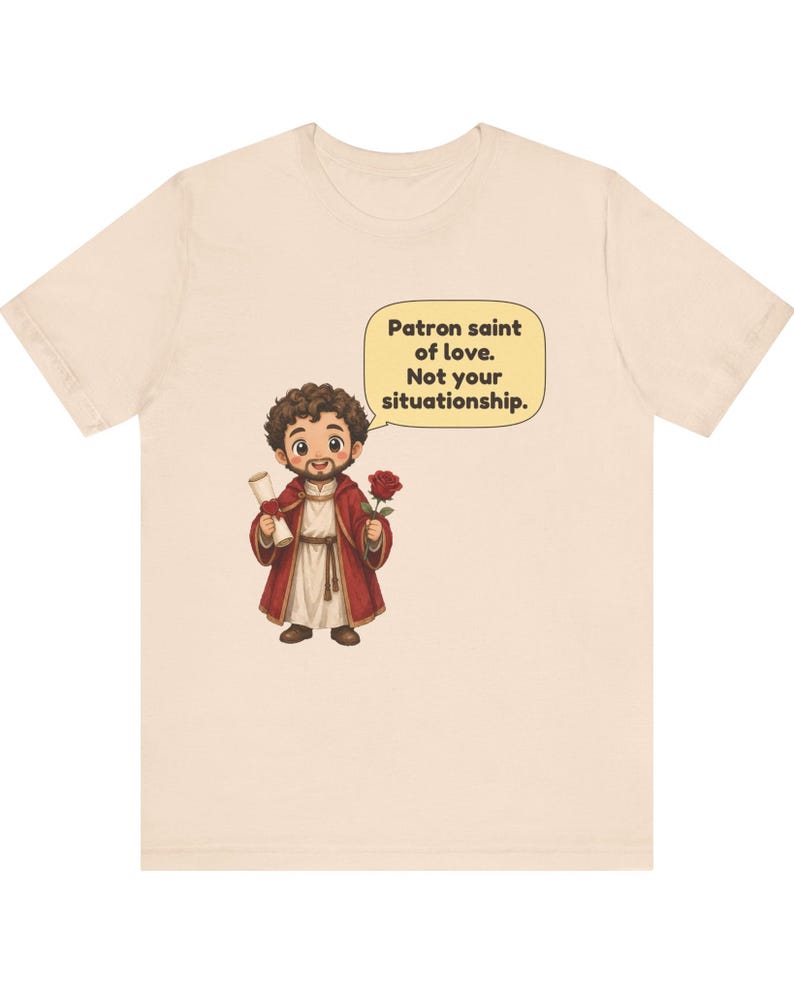Puede incluir: Una camiseta de color beige claro con una ilustraci&oacute;n de dibujos animados de una figura barbuda con t&uacute;nica roja que sostiene un pergamino y una rosa roja. Un globo de texto contiene: Patron saint of love. Not your situationship.