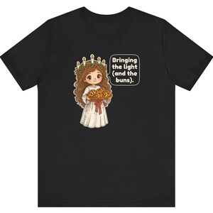 May include: A black t-shirt featuring a graphic of a character with long brown hair wearing a white robe and a crown of candles with greenery. The character holds a tray of buns and a speech bubble with the text: Bringing the light (and the buns).