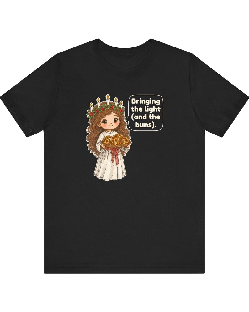 May include: A black t-shirt featuring a graphic of a character with long brown hair wearing a white robe and a crown of candles with greenery. The character holds a tray of buns and a speech bubble with the text: Bringing the light (and the buns).