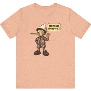 K&ouml;nnte beinhalten: Ein pfirsichfarbenes Kurzarm-T-Shirt mit einer Grafik einer Holzpuppe mit langer Nase, die einen Hut und ein gemustertes Outfit tr&auml;gt. Eine Sprechblase neben der Figur enth&auml;lt den Text, Honest! (Mostly.) in dunkelbraunen Buchstaben.