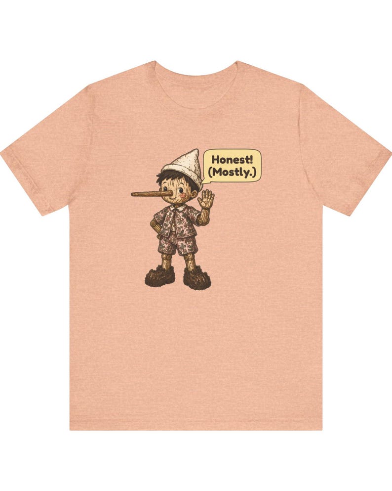 K&ouml;nnte beinhalten: Ein pfirsichfarbenes Kurzarm-T-Shirt mit einer Grafik einer Holzpuppe mit langer Nase, die einen Hut und ein gemustertes Outfit tr&auml;gt. Eine Sprechblase neben der Figur enth&auml;lt den Text, Honest! (Mostly.) in dunkelbraunen Buchstaben.