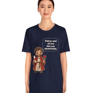 Puede incluir: Una camiseta azul marino de manga corta con una ilustraci&oacute;n de dibujos animados de una figura barbuda con t&uacute;nica roja que sostiene un pergamino y una rosa roja. Un globo de texto contiene: Patron saint of love. Not your situationship.