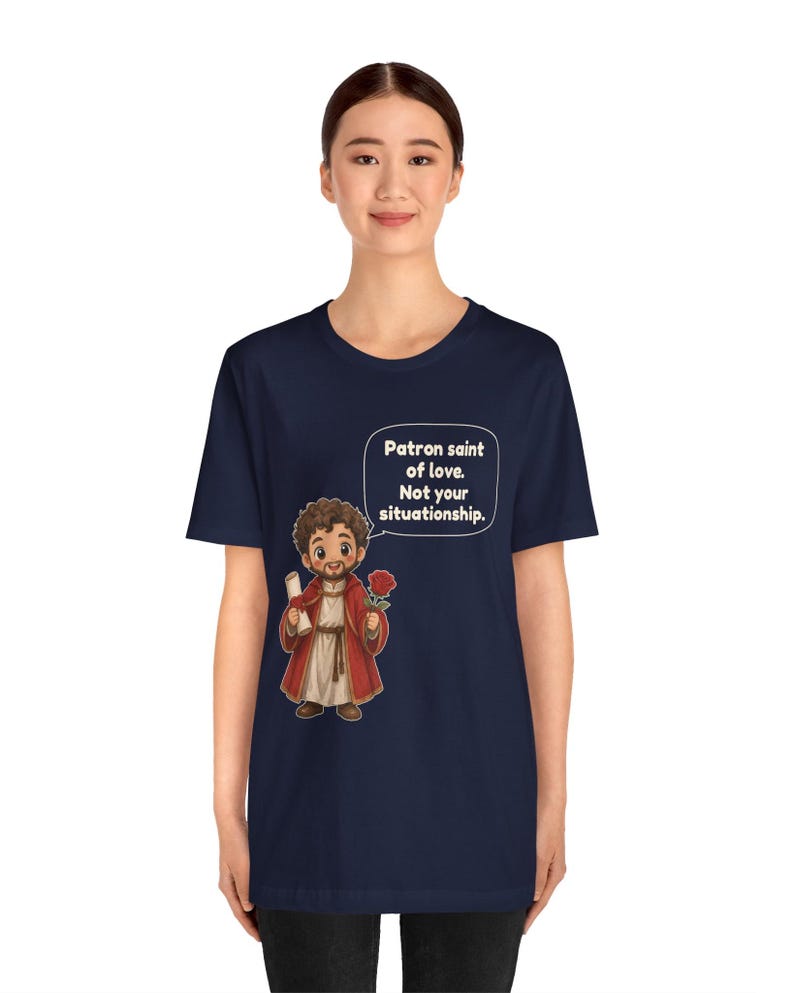 Puede incluir: Una camiseta azul marino de manga corta con una ilustraci&oacute;n de dibujos animados de una figura barbuda con t&uacute;nica roja que sostiene un pergamino y una rosa roja. Un globo de texto contiene: Patron saint of love. Not your situationship.