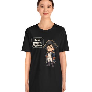 May include: A black t-shirt featuring a cartoon illustration of a historical figure in a blue military uniform and bicorne hat. A speech bubble next to the character contains the text: Small emperor. Big plans. The design is centered on the chest.