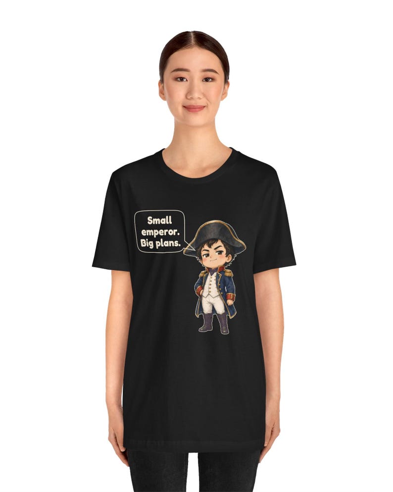 May include: A black t-shirt featuring a cartoon illustration of a historical figure in a blue military uniform and bicorne hat. A speech bubble next to the character contains the text: Small emperor. Big plans. The design is centered on the chest.