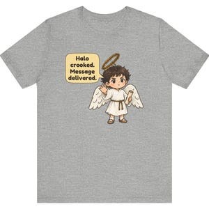 May include: A heather gray t-shirt featuring a cartoon angel with brown hair, white wings, and a crooked halo. The angel wears a white robe with a brown belt and holds a scroll. A speech bubble contains the text: Halo crooked. Message delivered.