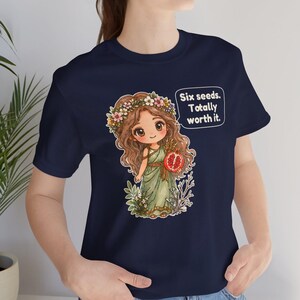 May include: A navy blue t-shirt featuring a graphic of a cartoon character with long brown hair, a flower crown, and a green dress, holding a pomegranate. A speech bubble next to the character contains the text: Six seeds. Totally worth it.