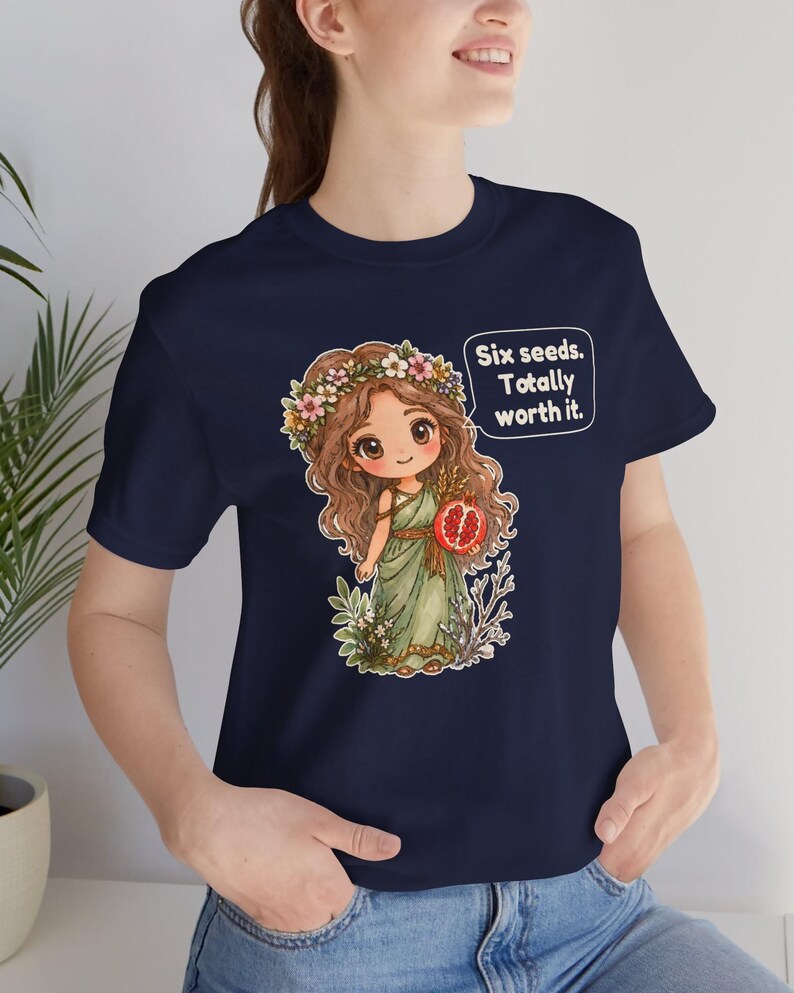 May include: A navy blue t-shirt featuring a graphic of a cartoon character with long brown hair, a flower crown, and a green dress, holding a pomegranate. A speech bubble next to the character contains the text: Six seeds. Totally worth it.