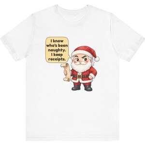 May include: A white t-shirt featuring a cartoon illustration of Santa Claus holding a scroll. A speech bubble next to him contains the text: I know who's been naughty. I keep receipts. Santa wears a red suit with white trim, a black belt, and black boots.