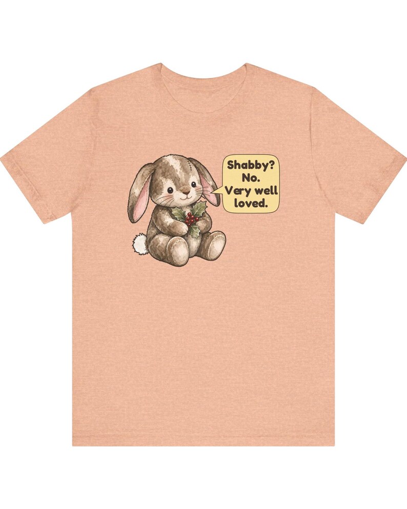 May include: A peach-colored short-sleeve t-shirt featuring a brown bunny illustration holding holly. A speech bubble next to the bunny contains the text: Shabby? No. Very well loved.