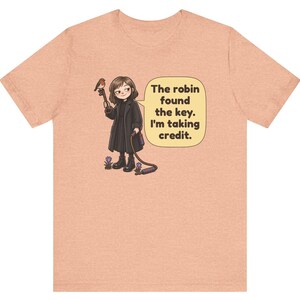 May include: A peach-colored t-shirt featuring a cartoon illustration of a person in a black coat holding a key with a robin perched on it. A speech bubble contains the text: The robin found the key. I'm taking credit. Small purple flowers are at the feet.