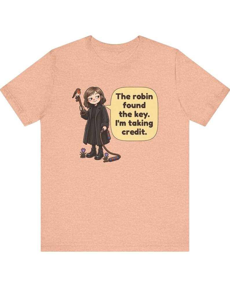 May include: A peach-colored t-shirt featuring a cartoon illustration of a person in a black coat holding a key with a robin perched on it. A speech bubble contains the text: The robin found the key. I'm taking credit. Small purple flowers are at the feet.
