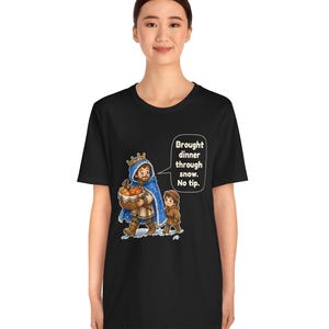May include: A black t-shirt featuring a graphic of a king in a blue cloak carrying a basket of bread, accompanied by a child. A speech bubble contains the text: Brought dinner through snow. No tip. The design is centered on the chest.
