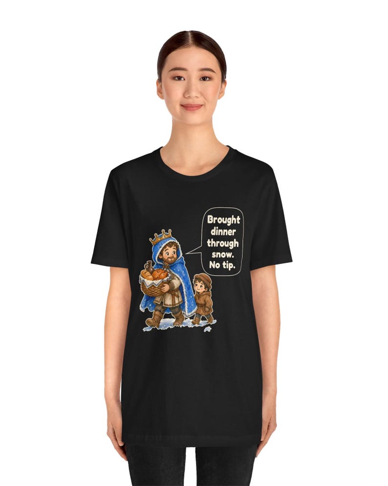 May include: A black t-shirt featuring a graphic of a king in a blue cloak carrying a basket of bread, accompanied by a child. A speech bubble contains the text: Brought dinner through snow. No tip. The design is centered on the chest.
