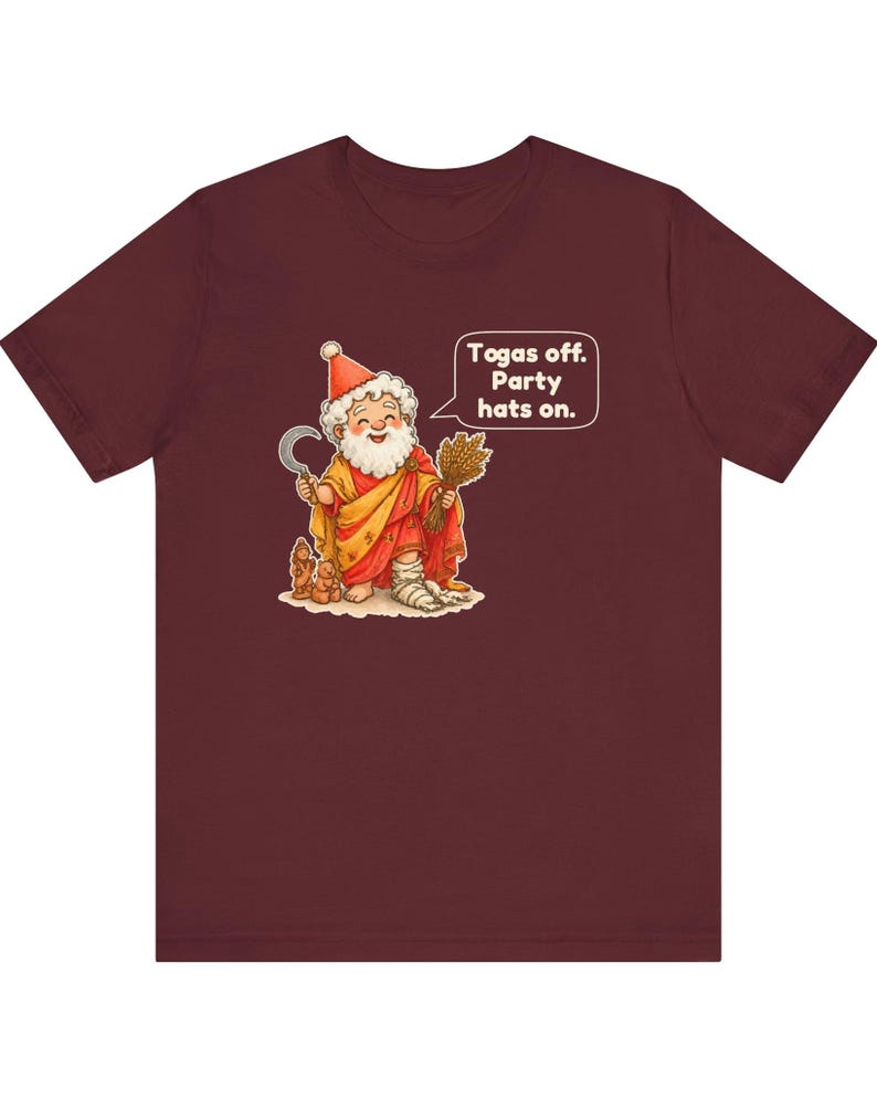 May include: A maroon t-shirt featuring a cartoon illustration of a bearded figure wearing a red party hat and a yellow and red robe. The figure holds a sickle and a bundle of wheat, with a speech bubble that reads: Togas off. Party hats on.