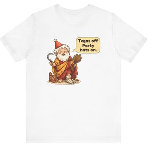 May include: A white t-shirt featuring a cartoon illustration of a bearded figure wearing a red and yellow robe, a red party hat, and a bandage on one foot. The figure holds a sickle and wheat, with a speech bubble that reads: Togas off. Party hats on.