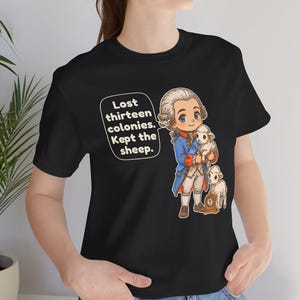 Peut inclure: Un t-shirt noir avec une illustration de style dessin anim&eacute; repr&eacute;sentant un personnage historique en manteau bleu tenant un petit mouton, avec un autre mouton et une horloge. Une bulle contient le texte : Lost thirteen colonies. Kept the sheep.