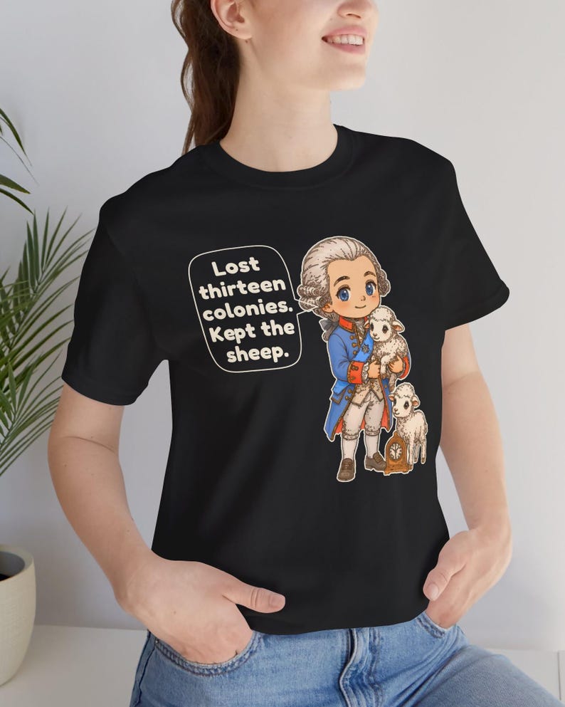 Peut inclure: Un t-shirt noir avec une illustration de style dessin anim&eacute; repr&eacute;sentant un personnage historique en manteau bleu tenant un petit mouton, avec un autre mouton et une horloge. Une bulle contient le texte : Lost thirteen colonies. Kept the sheep.