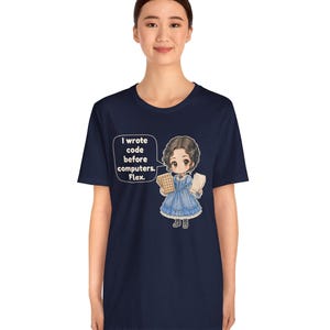 May include: A navy blue t-shirt featuring a graphic of a person in a blue Victorian-style dress holding punch cards. A speech bubble next to the character contains the text: I wrote code before computers. Flex.