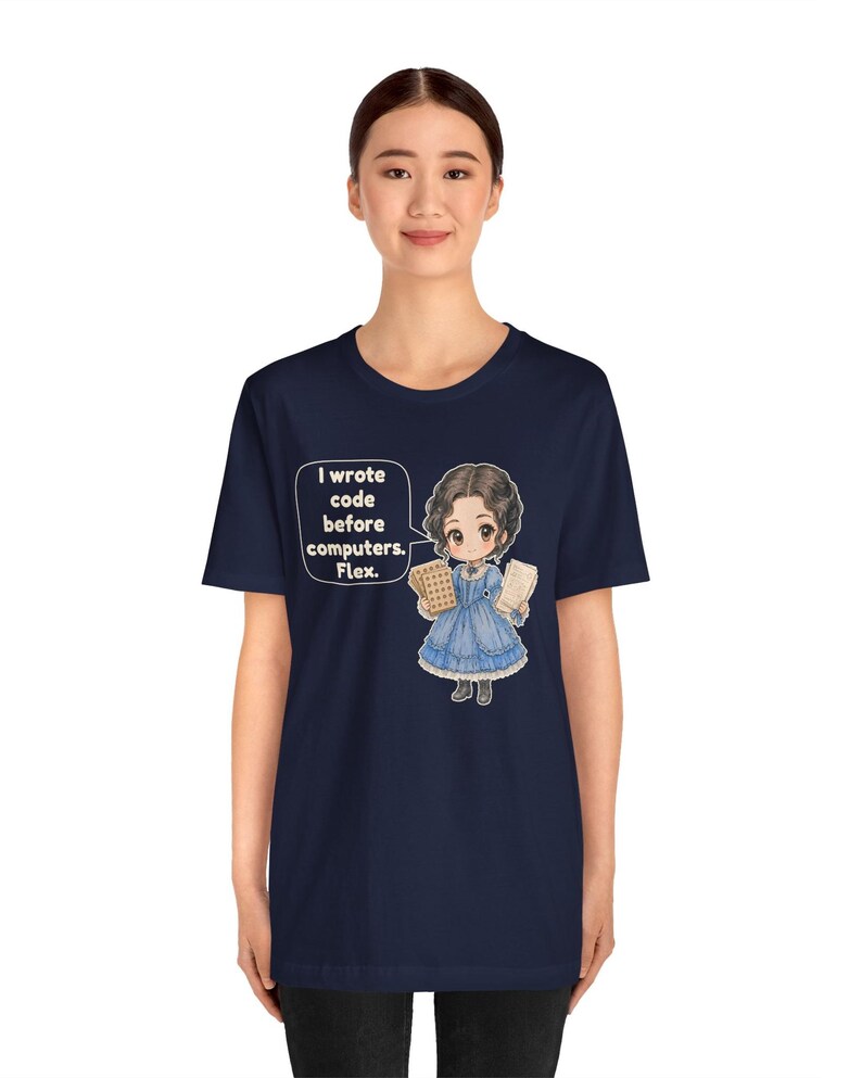 May include: A navy blue t-shirt featuring a graphic of a person in a blue Victorian-style dress holding punch cards. A speech bubble next to the character contains the text: I wrote code before computers. Flex.